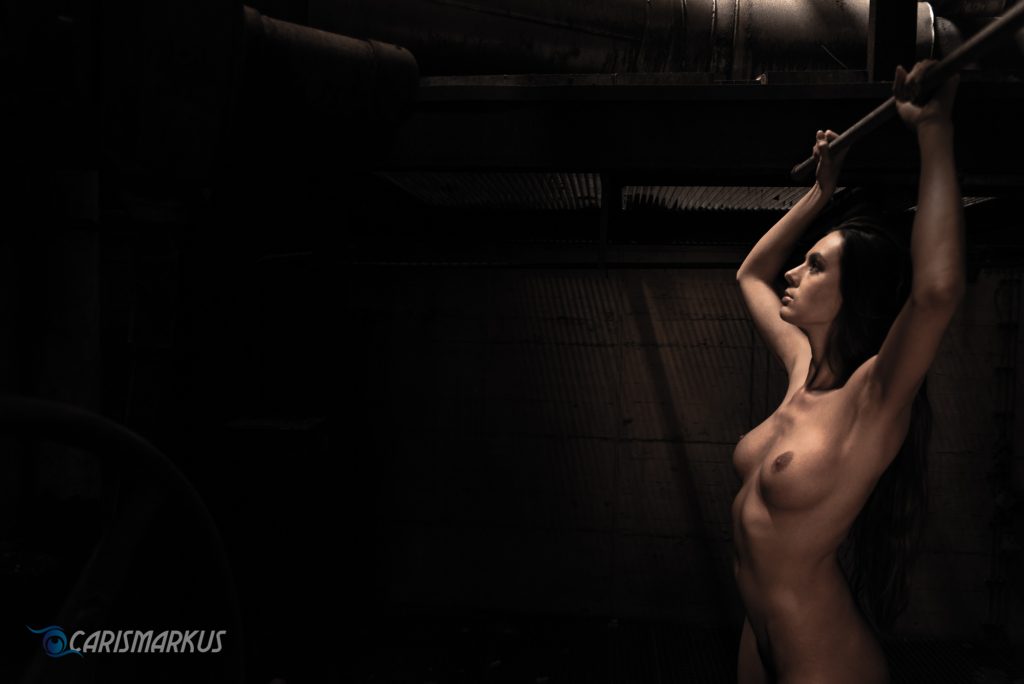 Topmodel Nikolart in harsh contrast between darkness, shadows and a single reay of light coming from above and enlighting her beautiful body in the rough industrial environment.